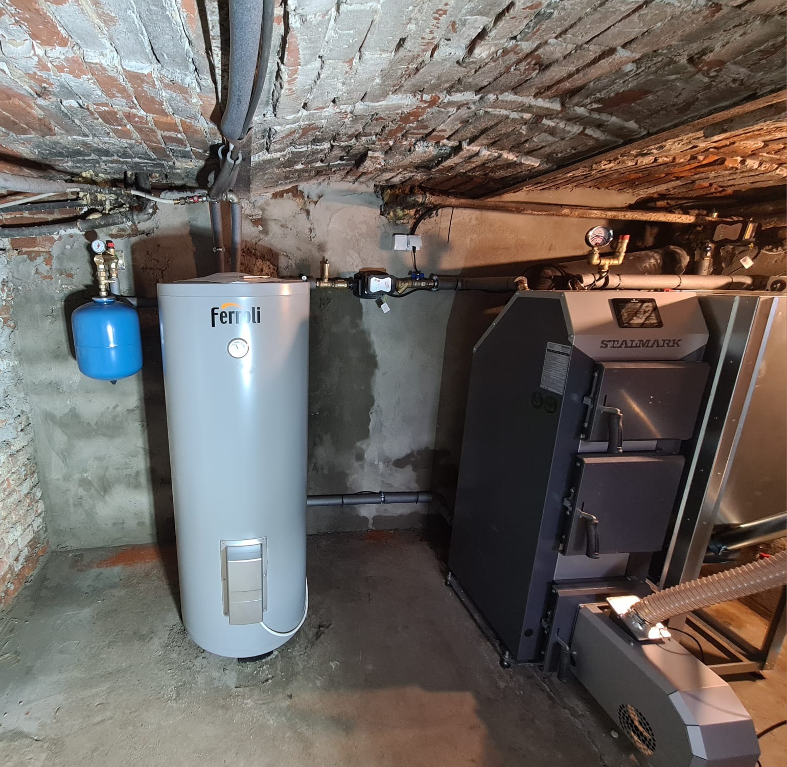 Kotłownia z piecem na paliwo stałe Stalmark i zasobnikiem ciepłej wody Ferroli, widoczne instalacje, ceglany sufit piwnicy.