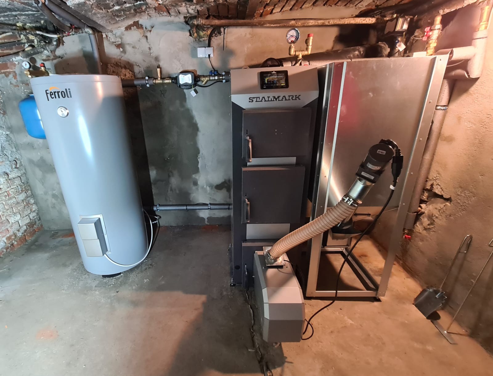 Nowoczesny piec na paliwo stałe Stalmark z podajnikiem ślimakowym i zasobnikiem na wodę Ferroli w kotłowni o surowym wykończeniu ścian.