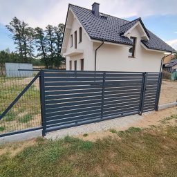 Manufaktura Spawalnictwa Adam Grześkowiak - Nowoczesne, poziome przęsła w kolorze grafitowym, tworzące ogrodzenie posesji z domem w tle. Widoczny fragment siatki ogrodzeniowej i betonowa podmurówka.