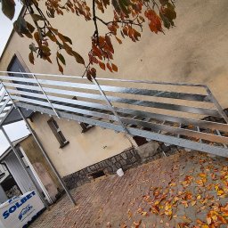 Manufaktura Spawalnictwa Adam Grześkowiak - Zewnętrzne, metalowe schody z balustradą, prowadzące do budynku. Widoczne jesienne liście na brukowanym podłożu i fragment elewacji. Ujęcie z dołu, pod kątem.