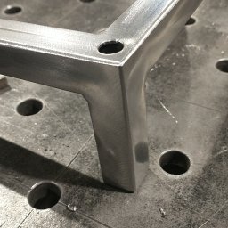 Manufaktura Spawalnictwa Adam Grześkowiak - Metalowy element konstrukcyjny z otworem, leżący na perforowanym stole warsztatowym. Widoczne ślady obróbki i drobne opiłki.