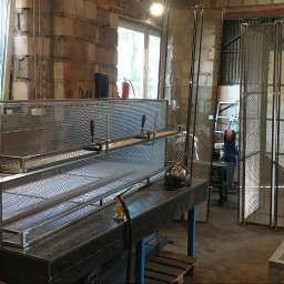 Manufaktura Spawalnictwa Adam Grześkowiak - Warsztatowa sceneria z metalowymi konstrukcjami w trakcie tworzenia. Widoczny stół roboczy, elementy siatkowe i narzędzia. Surowy, industrialny klimat.