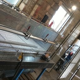 Manufaktura Spawalnictwa Adam Grześkowiak - Warsztat ślusarski: metalowa konstrukcja z siatki na stole spawalniczym, kaski i narzędzia w tle, surowe mury z cegły i okno z jasnym światłem.