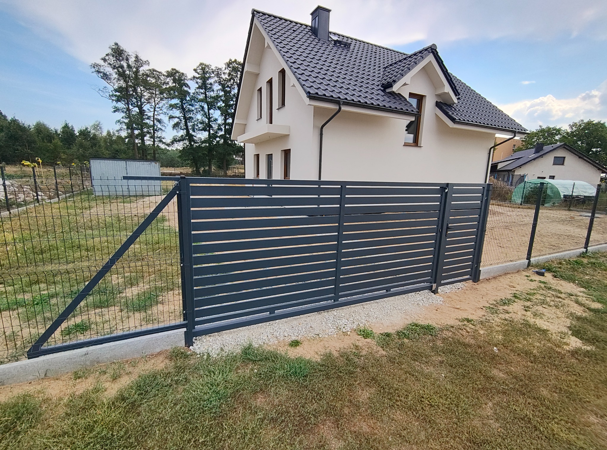 Nowoczesne, poziome przęsła w kolorze grafitowym, tworzące ogrodzenie posesji z domem w tle. Widoczny fragment siatki ogrodzeniowej i betonowa podmurówka.