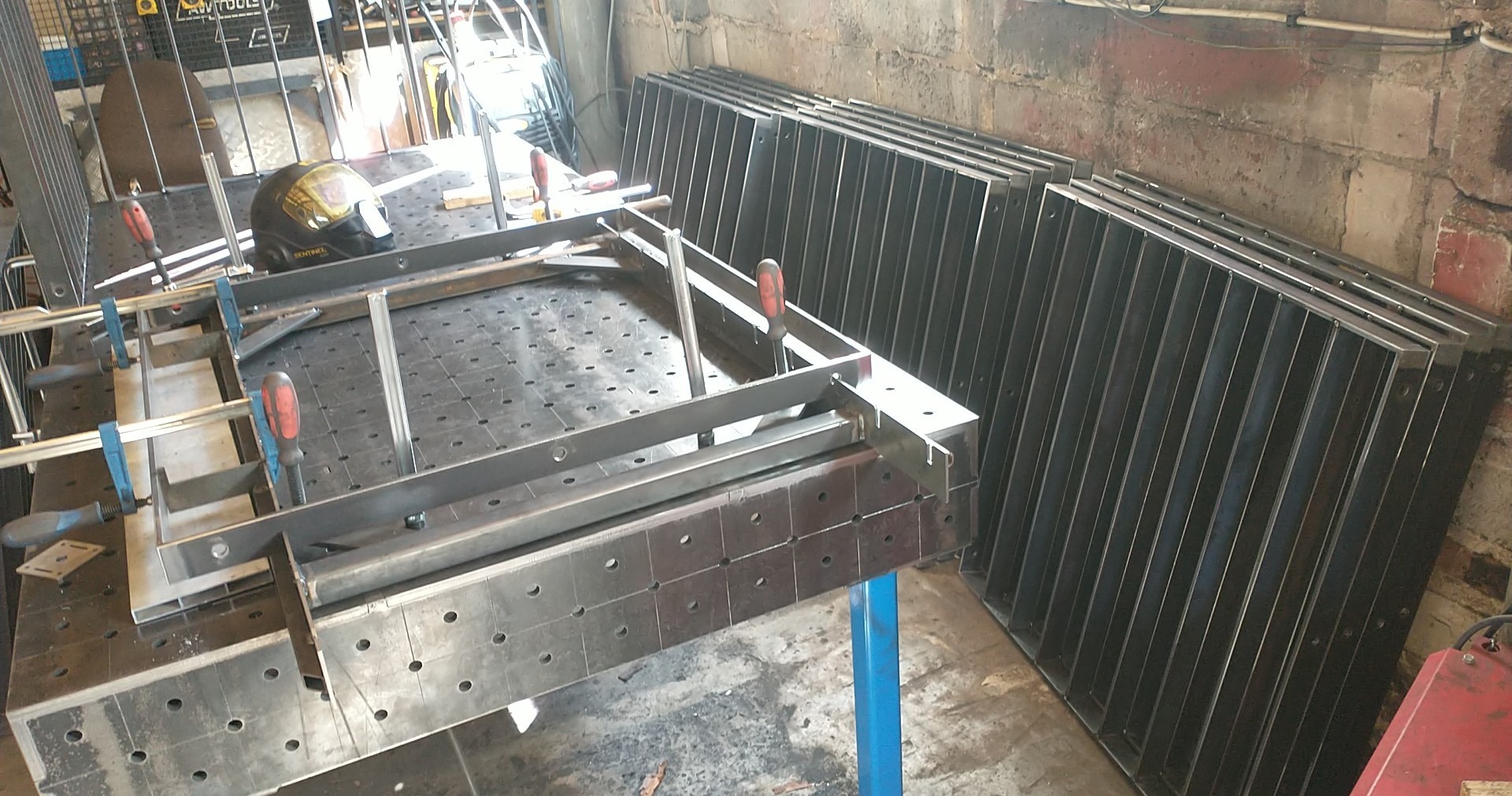 Metalowa konstrukcja w trakcie montażu na perforowanym stole warsztatowym, obok gotowe elementy. Widoczny kask spawalniczy i narzędzia.