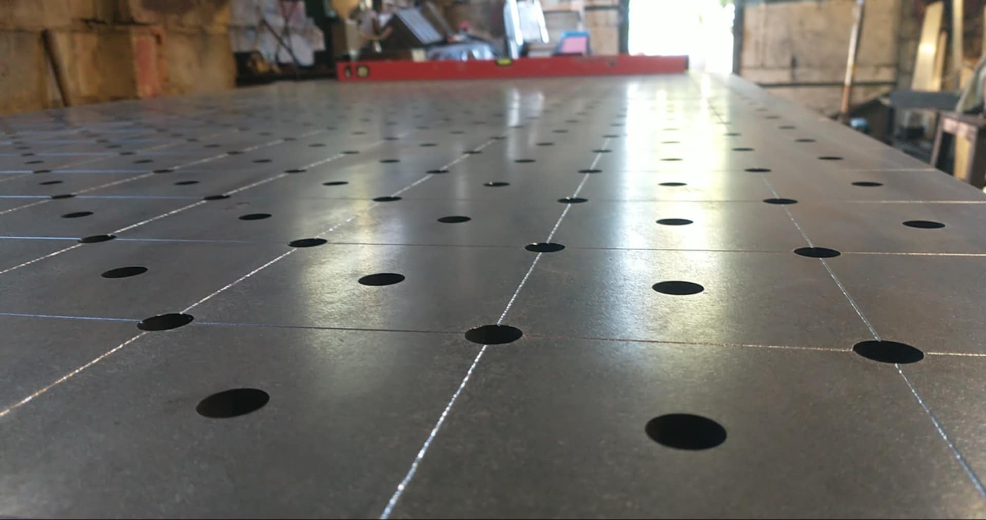 Stół spawalniczy z otworami i poziomnicą w tle, ukazujący precyzję wykonania i gotowość do pracy. Metalowa konstrukcja z regularnym układem otworów.