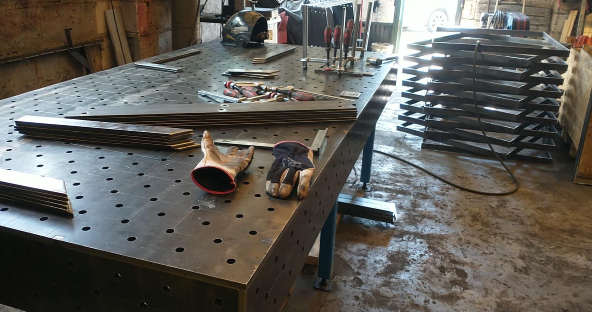 Warsztatowa przestrzeń robocza z perforowanym stołem, narzędziami, rękawicami i stosem metalowych elementów, sugerująca etap obróbki metalu.