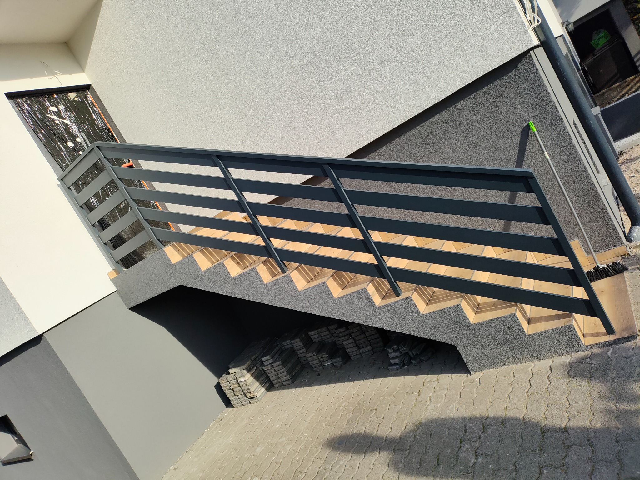 Zewnętrzne schody z metalową balustradą, prowadzące do budynku o nowoczesnej architekturze. Widoczne detale wykończenia i ułożona kostka brukowa.