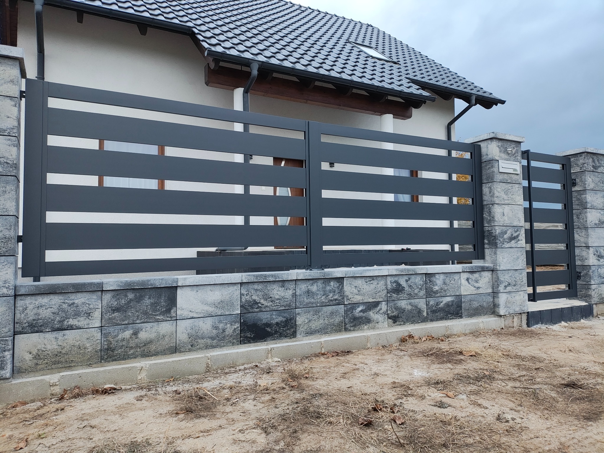Nowoczesna, pozioma brama wjazdowa i furtka z szarego metalu, osadzone na słupkach z szarej kostki. Tło stanowi dom z ciemnym dachem i jasną elewacją.