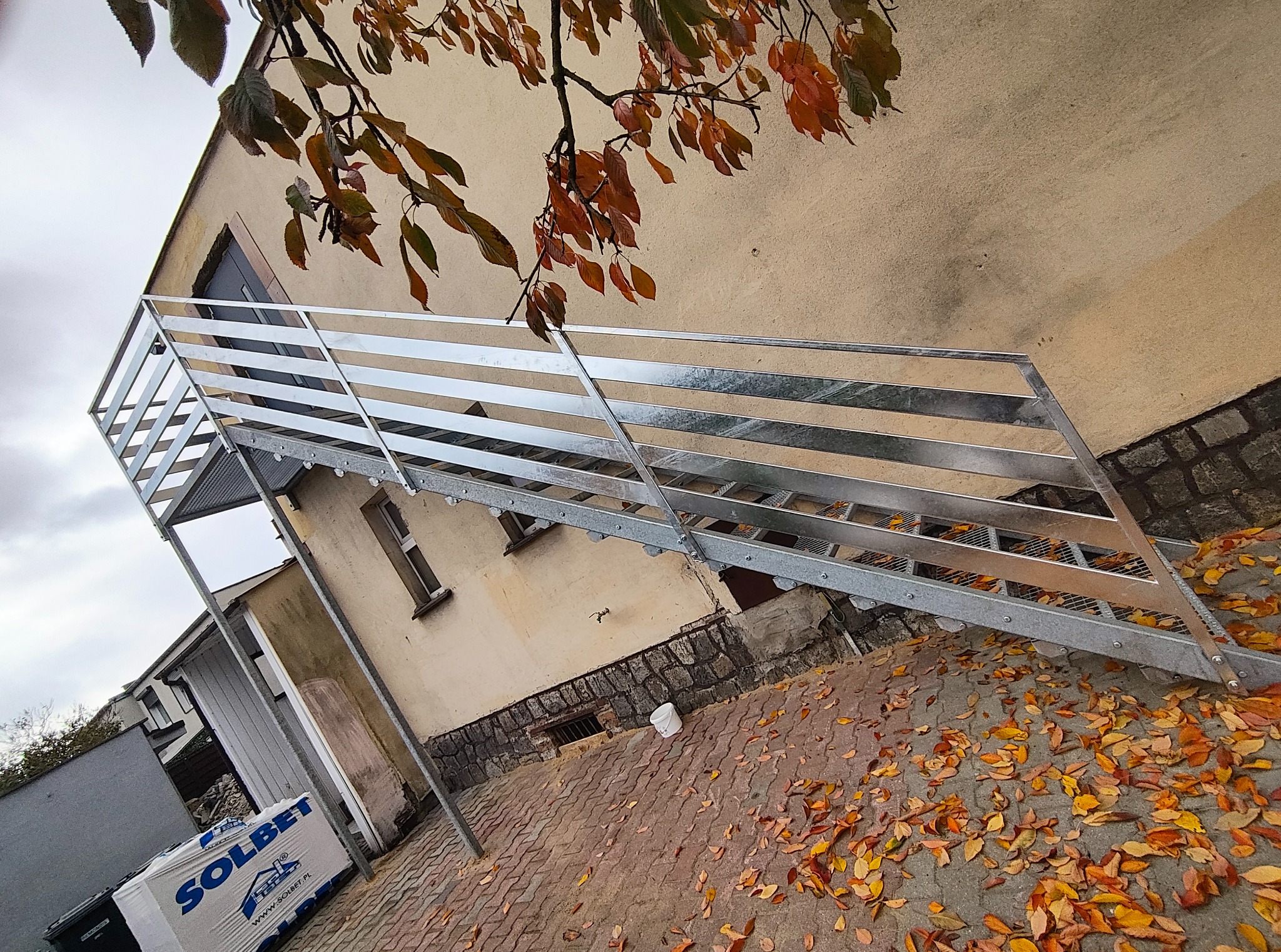 Zewnętrzne, metalowe schody z balustradą, prowadzące do budynku. Widoczne jesienne liście na brukowanym podłożu i fragment elewacji. Ujęcie z dołu, pod kątem.
