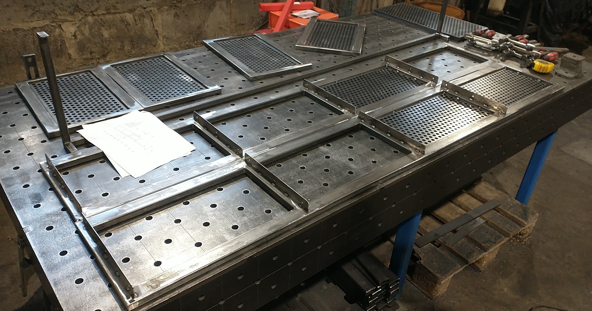 Metalowe ramy i perforowane płyty na stole spawalniczym z otworami, widoczne narzędzia i rysunek techniczny. Elementy przygotowane do dalszej obróbki.