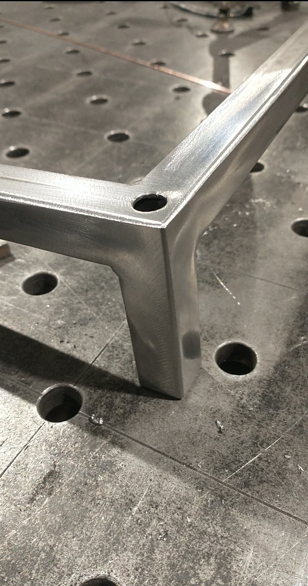 Metalowy element konstrukcyjny z otworem, leżący na perforowanym stole warsztatowym. Widoczne ślady obróbki i drobne opiłki.