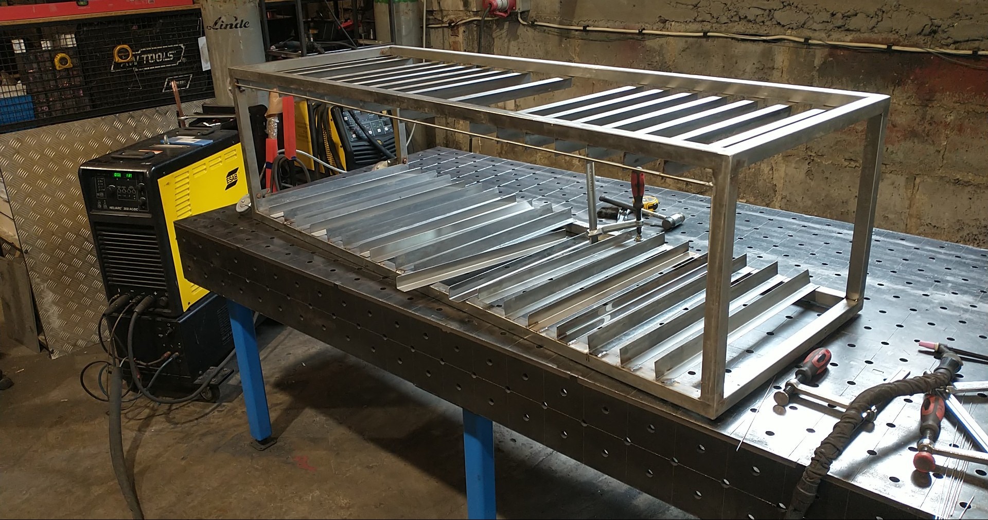 Spawanie konstrukcji metalowej z profili na stole spawalniczym. Widoczna spawarka ESAB. Elementy konstrukcji połączone klamrami. Surowy, warsztatowy klimat.