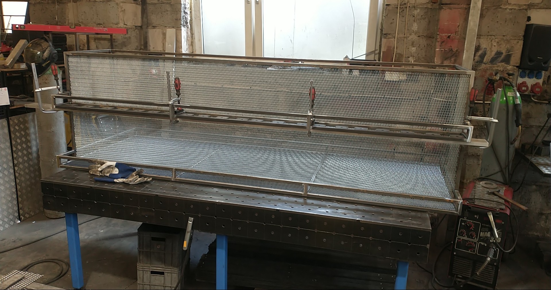 Konstrukcja z metalowej siatki na stole warsztatowym z narzędziami, w tle ceglana ściana i butla z gazem. Elementy mocowane za pomocą zacisków śrubowych.