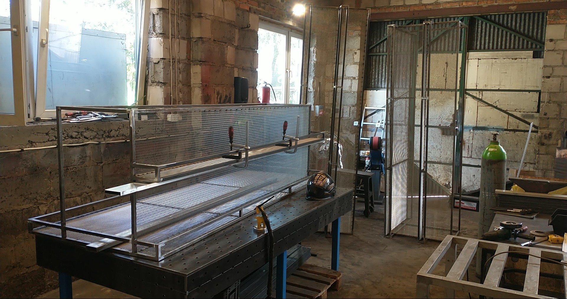 Warsztatowa sceneria z metalowymi konstrukcjami w trakcie tworzenia. Widoczny stół roboczy, elementy siatkowe i narzędzia. Surowy, industrialny klimat.
