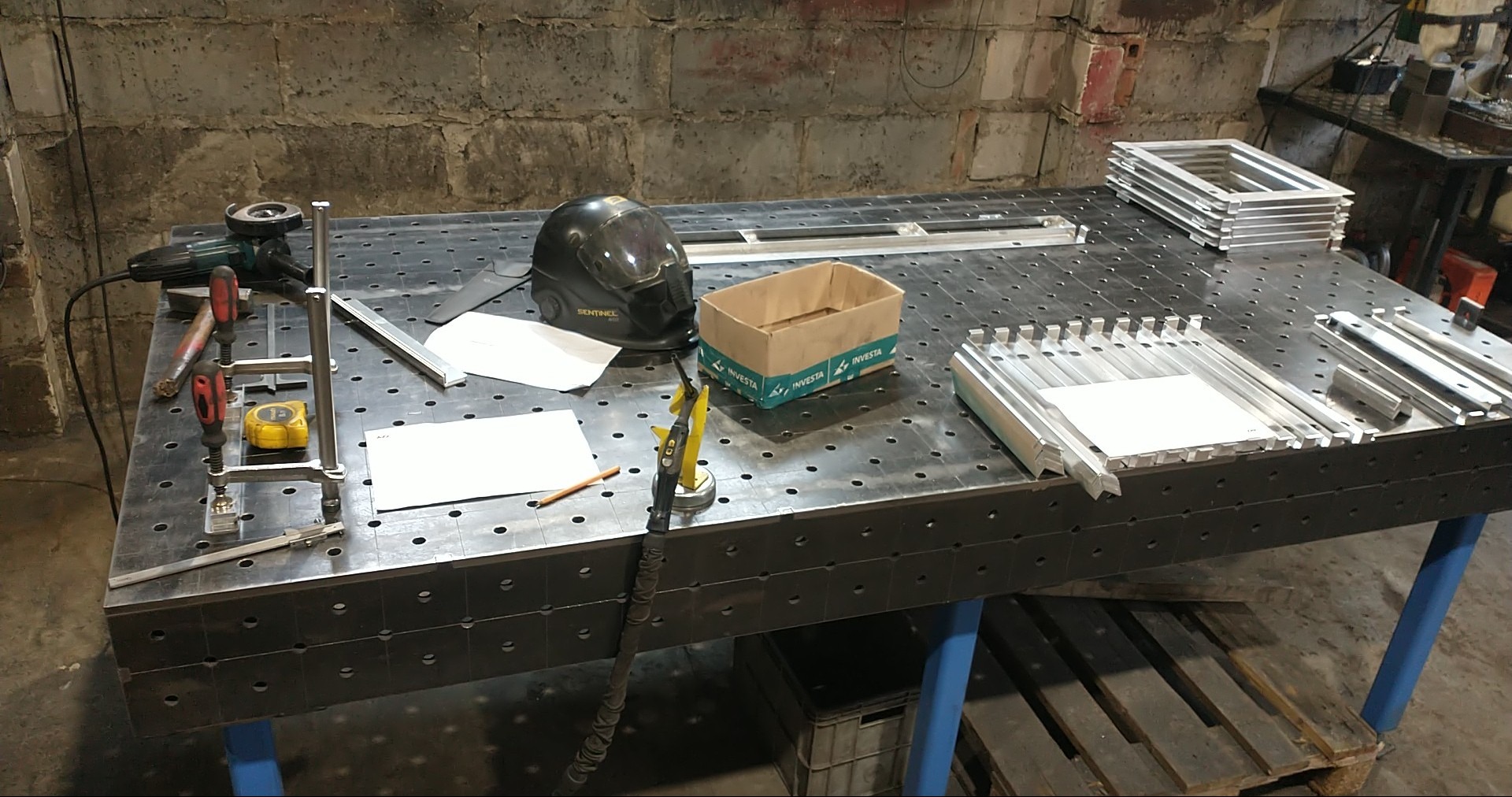 Stół roboczy z oprzyrządowaniem i elementami metalowymi w warsztacie w Opalenicy. Widoczny uchwyt spawalniczy, szlifierka kątowa i kask ochronny.