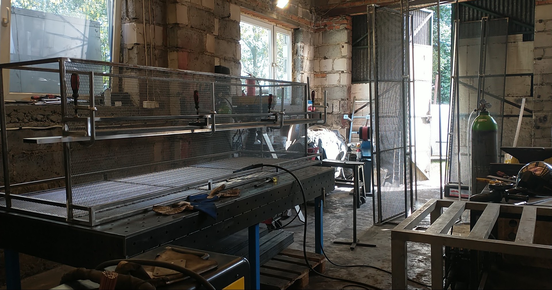 Metalowa konstrukcja z siatki w warsztacie, widoczne narzędzia i butla z gazem. Surowe wnętrze z ceglanymi ścianami i oknem w tle. Elementy w trakcie produkcji.