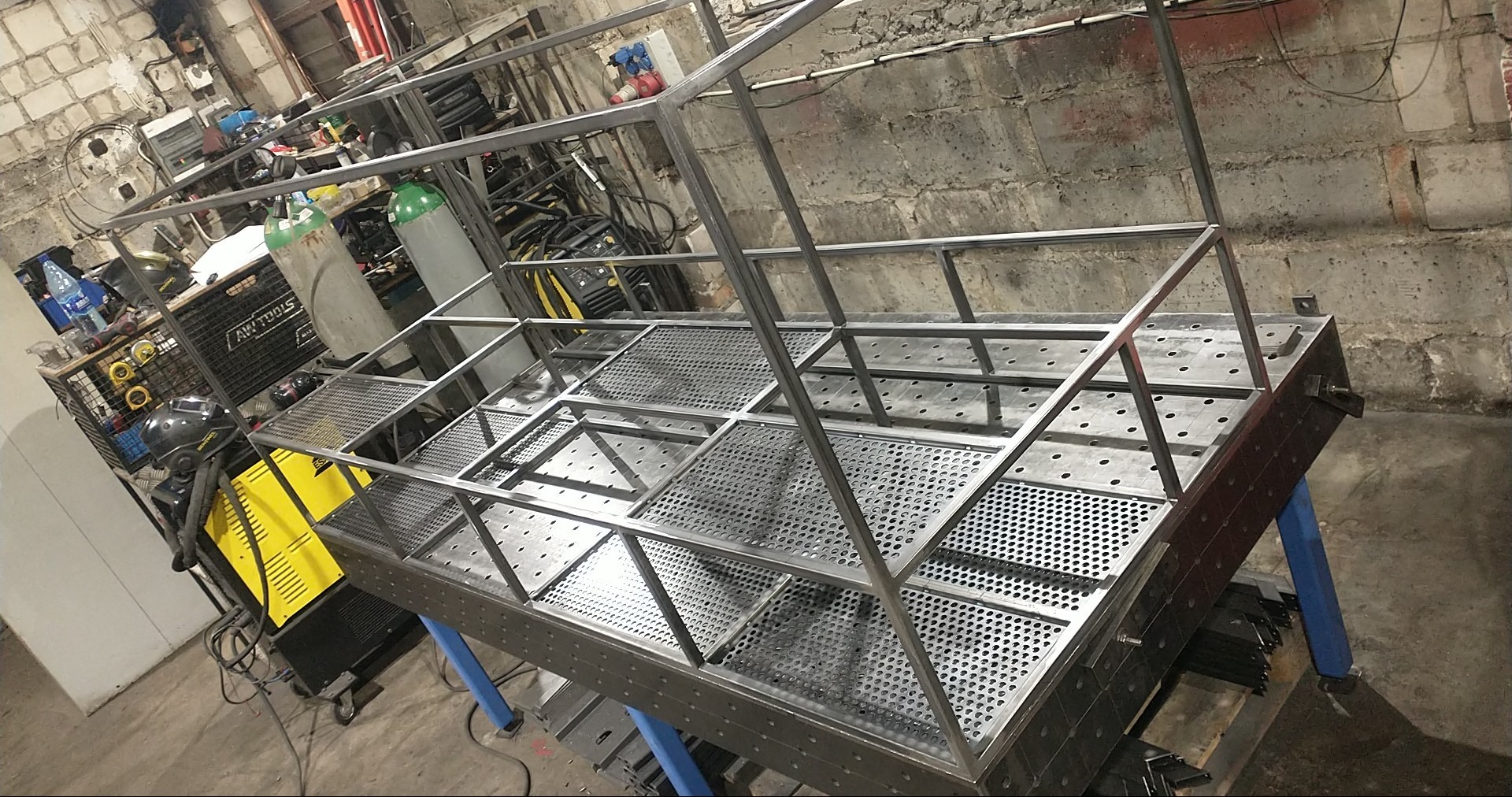 Metalowa konstrukcja z perforowanymi elementami, spawana na stole warsztatowym. W tle widoczny sprzęt spawalniczy i narzędzia w warsztacie.