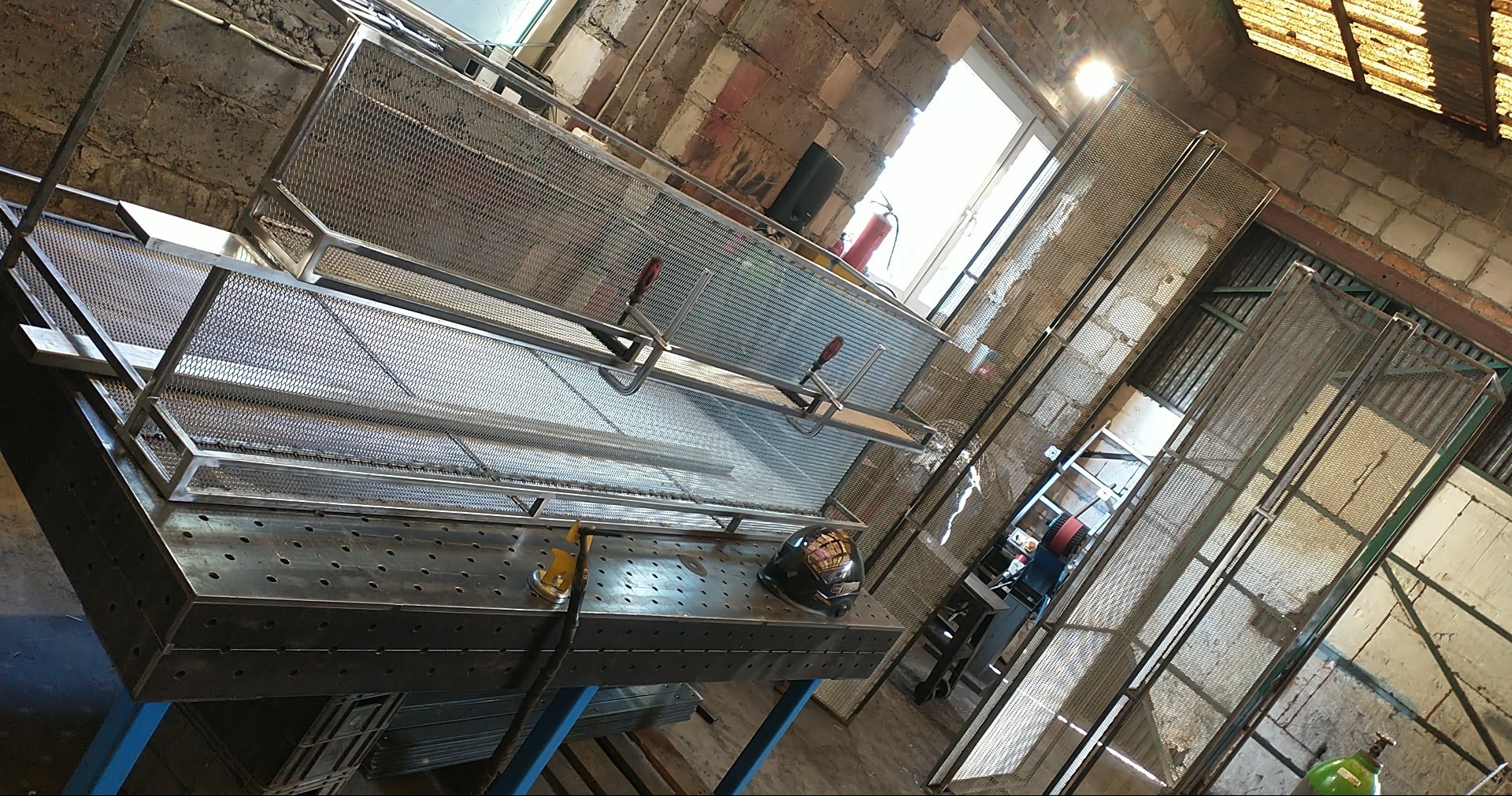 Warsztat ślusarski: metalowa konstrukcja z siatki na stole spawalniczym, kaski i narzędzia w tle, surowe mury z cegły i okno z jasnym światłem.