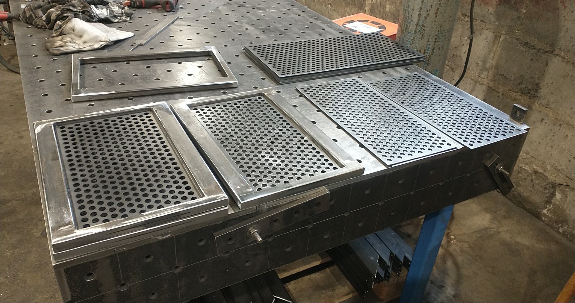 Metalowe ramy z perforowanymi wypełnieniami na stole warsztatowym. Widoczne ślady obróbki i spawania. Elementy przygotowane do dalszego montażu lub wykończenia.