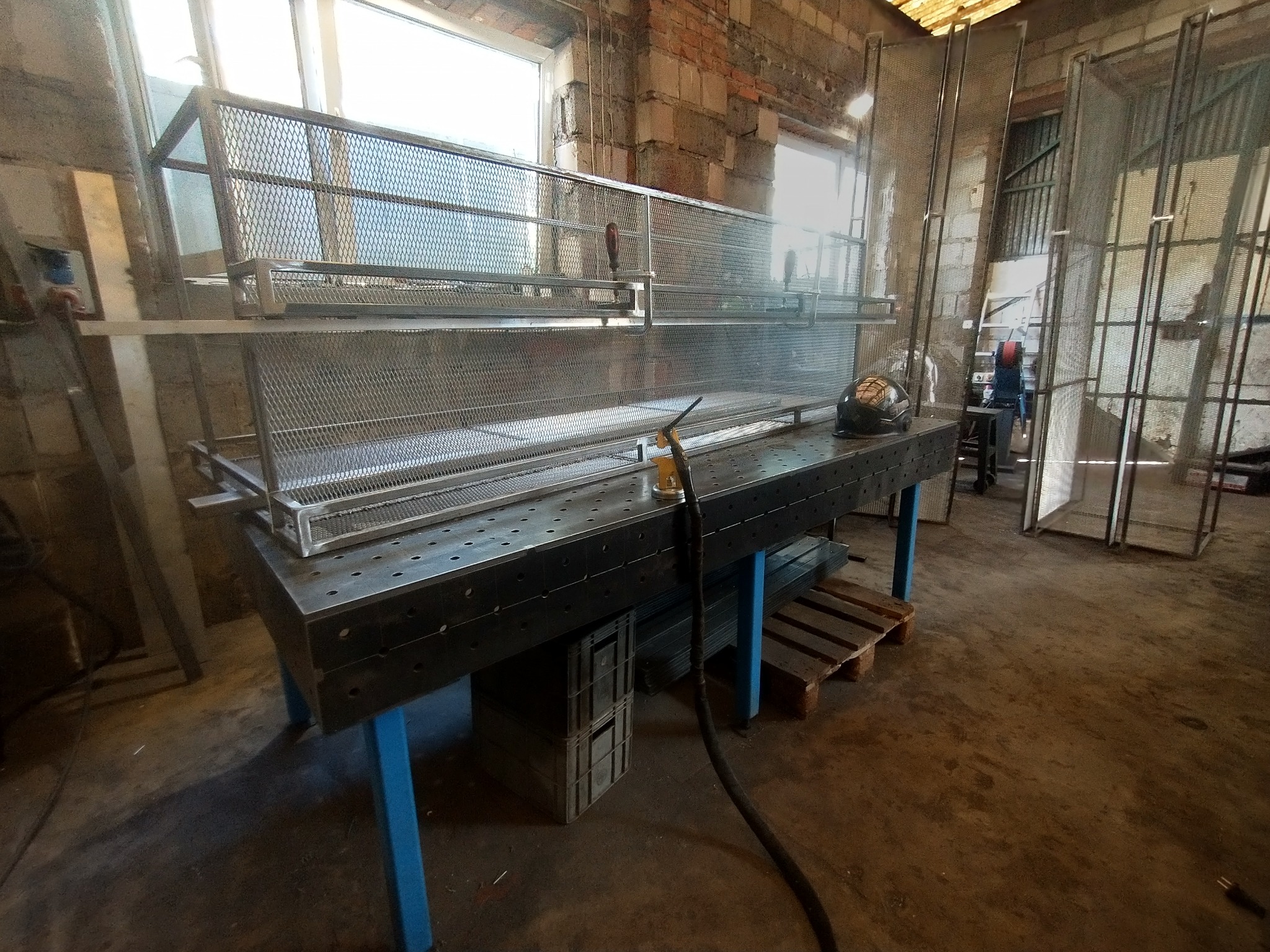 Metalowa konstrukcja z siatki na stole warsztatowym w hali, widoczny hełm spawalniczy i narzędzia. W tle okna i ceglana ściana. Surowy, industrialny klimat.