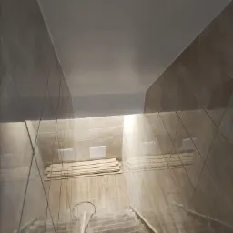 Klatka schodowa w trakcie wykańczania, wyłożona beżowymi płytkami, z widocznymi schodami i ułożonymi deskami na podłodze.