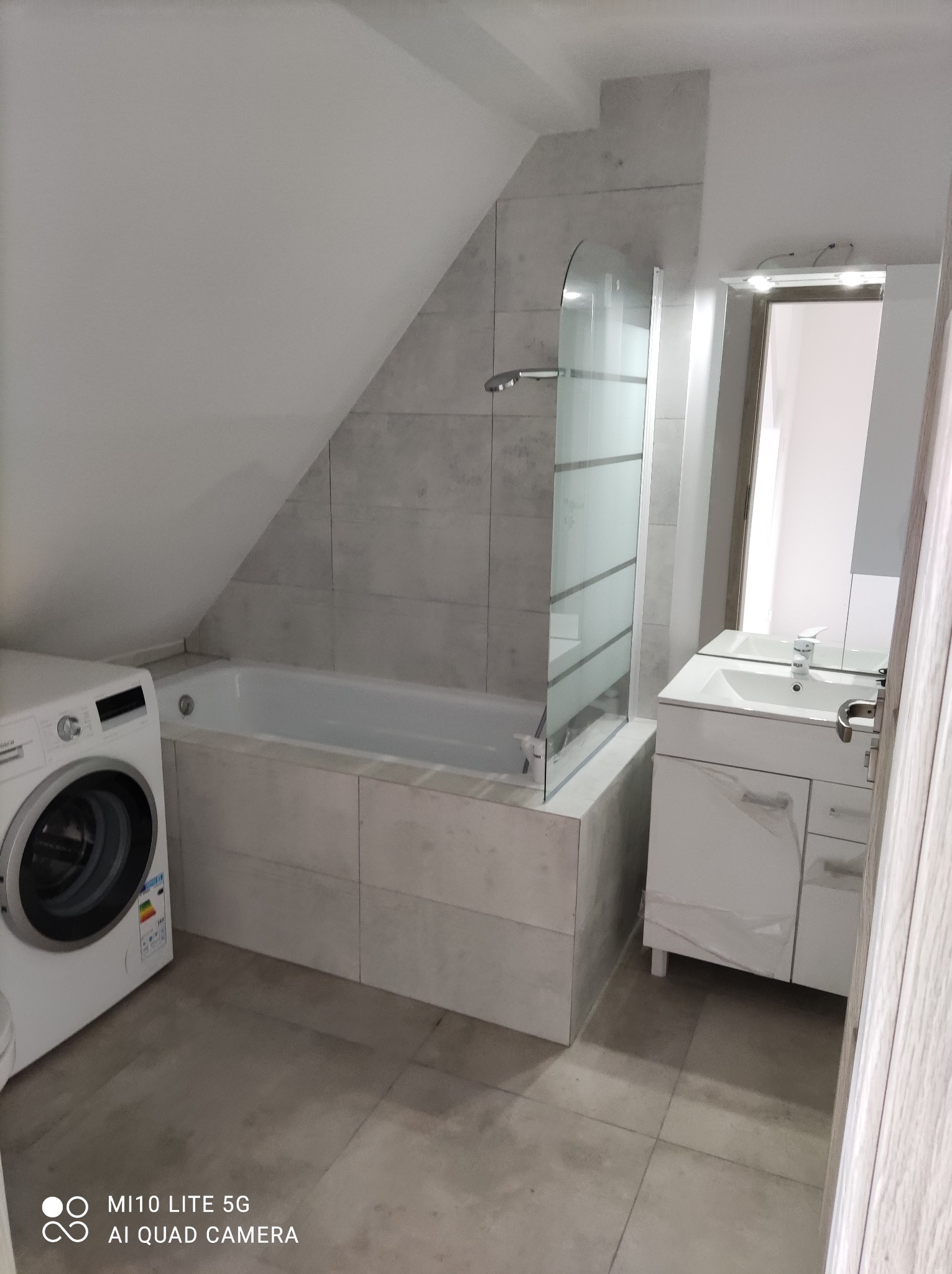 Szara łazienka na poddaszu z wanną obudowaną płytkami, kabiną prysznicową z częściowo matowego szkła, białą umywalką z szafką i pralką.