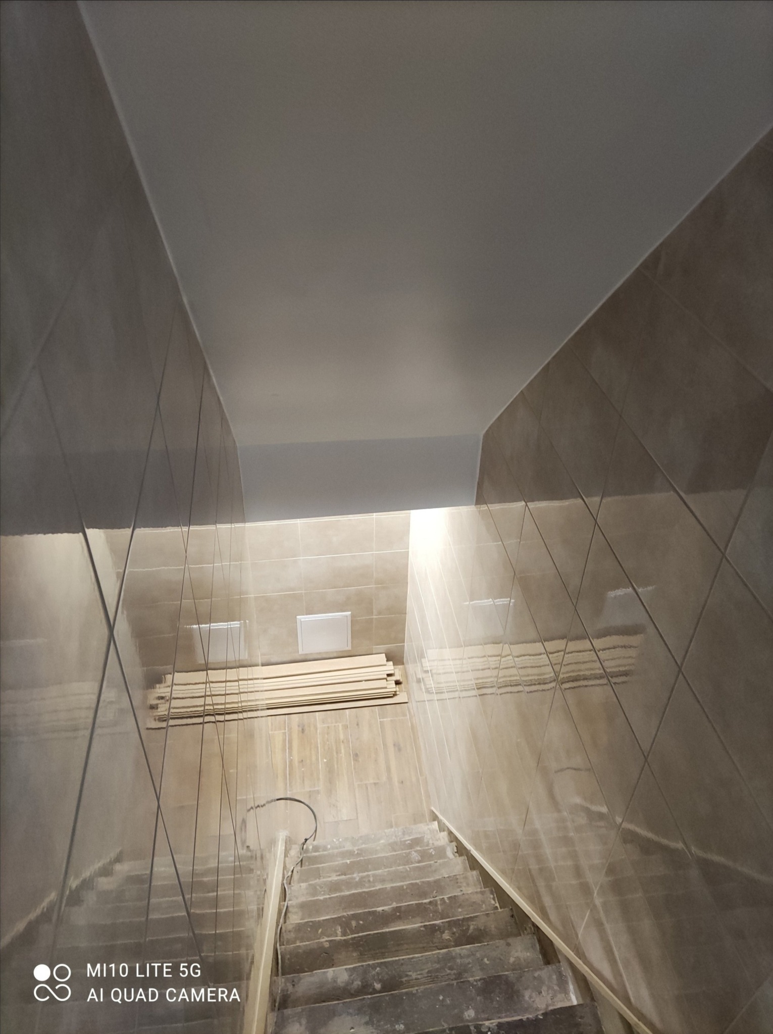 Klatka schodowa w trakcie wykańczania, wyłożona beżowymi płytkami, z widocznymi schodami i ułożonymi deskami na podłodze.