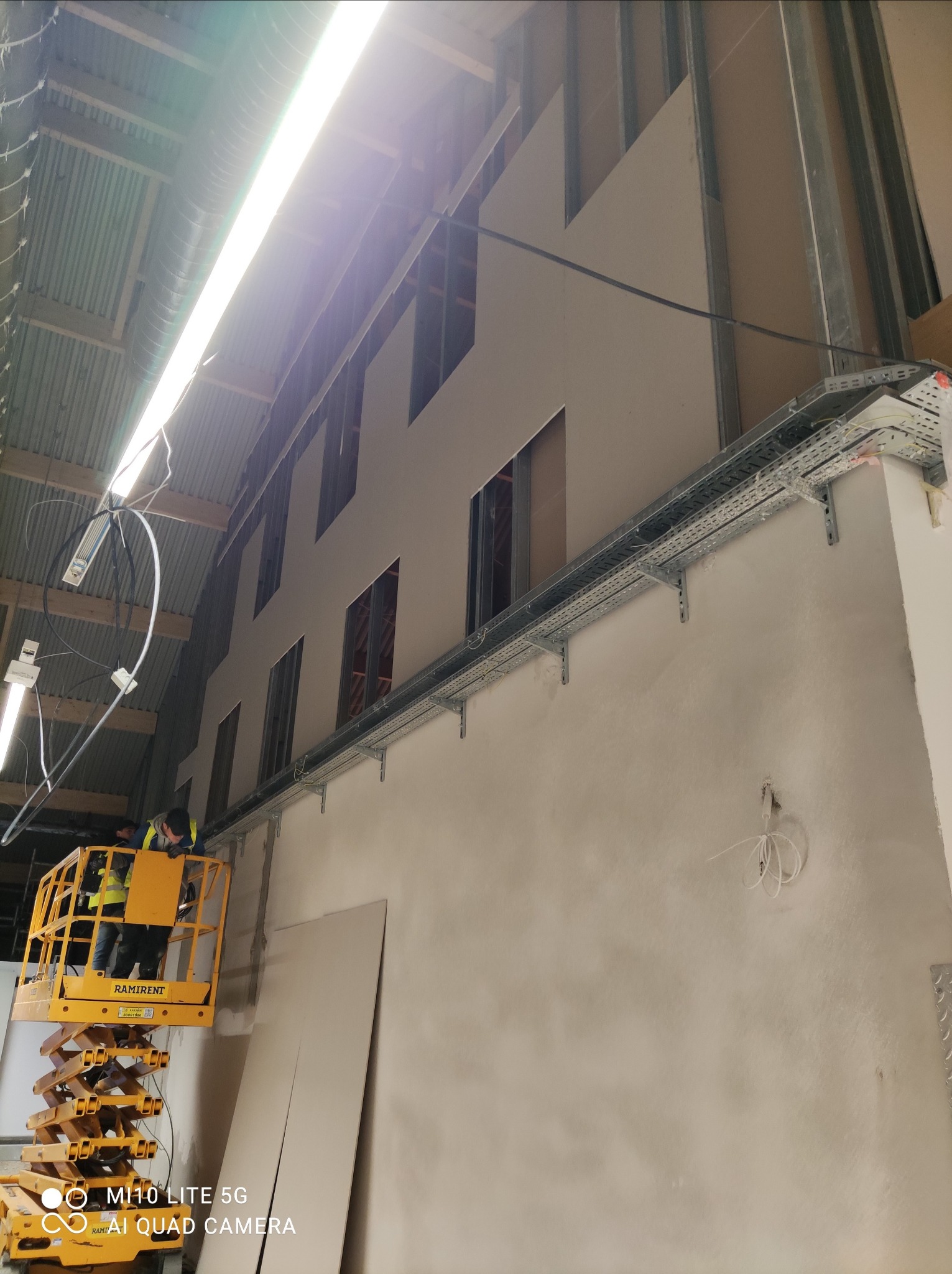 Montaż płyt gipsowo-kartonowych na ścianie w wysokim pomieszczeniu z użyciem podnośnika nożycowego Ramirent, widoczne profile konstrukcyjne i koryta kablowe pod sufitem.