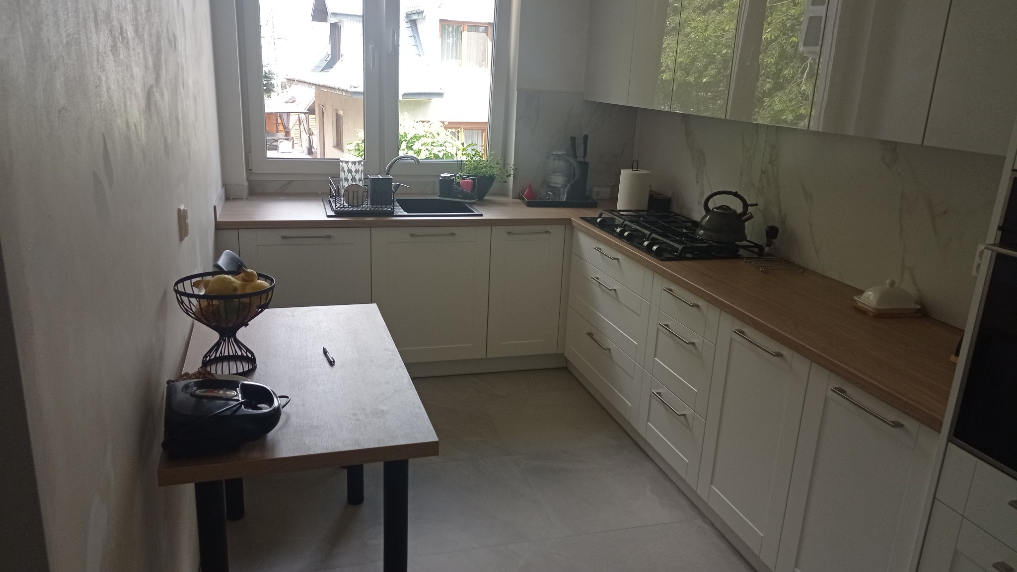 Jasna kuchnia z drewnianym blatem, widok na zewnątrz przez okno, na stole misa z owocami i czarny ekspres do kawy.