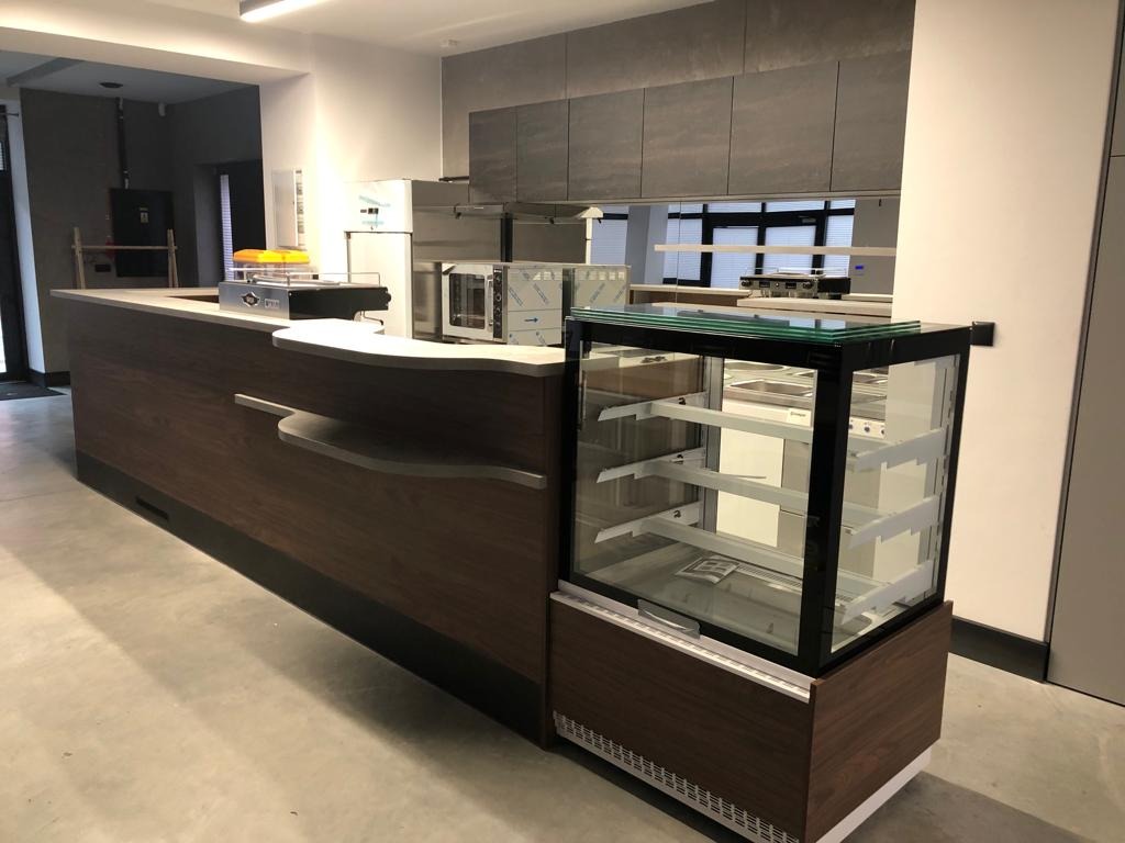 Nowoczesna lada barowa z ciemnego drewna z blatem i szklaną witryną chłodniczą w minimalistycznym wnętrzu lokalu gastronomicznego.