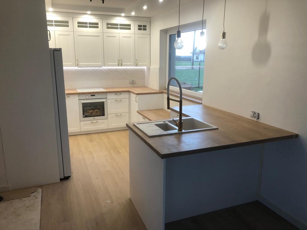 Jasna kuchnia z drewnianym blatem, wyspą kuchenną z dwukomorowym zlewem i baterią w kolorze mosiądzu, widok na okno z podmiejskim krajobrazem.