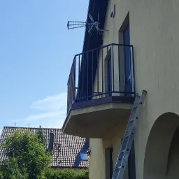 Metalowa balustrada balkonowa w kolorze czarnym, zamontowana na balkonie w budynku o beżowej elewacji, z widoczną drabiną opartą o ścianę i anteną na dachu.