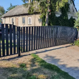 Nowoczesna, czarna brama wjazdowa z pionowych profili, z częściowo otwartym skrzydłem, na tle starego domu z oknem zasłoniętym reklamą.