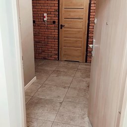 Rqbsqn Robert - Wykończony korytarz z ceglaną ścianą, drewnianymi drzwiami wewnętrznymi z czarną ościeżnicą i szarą podłogą z płytek, widok z otwartych drzwi wejściowych.