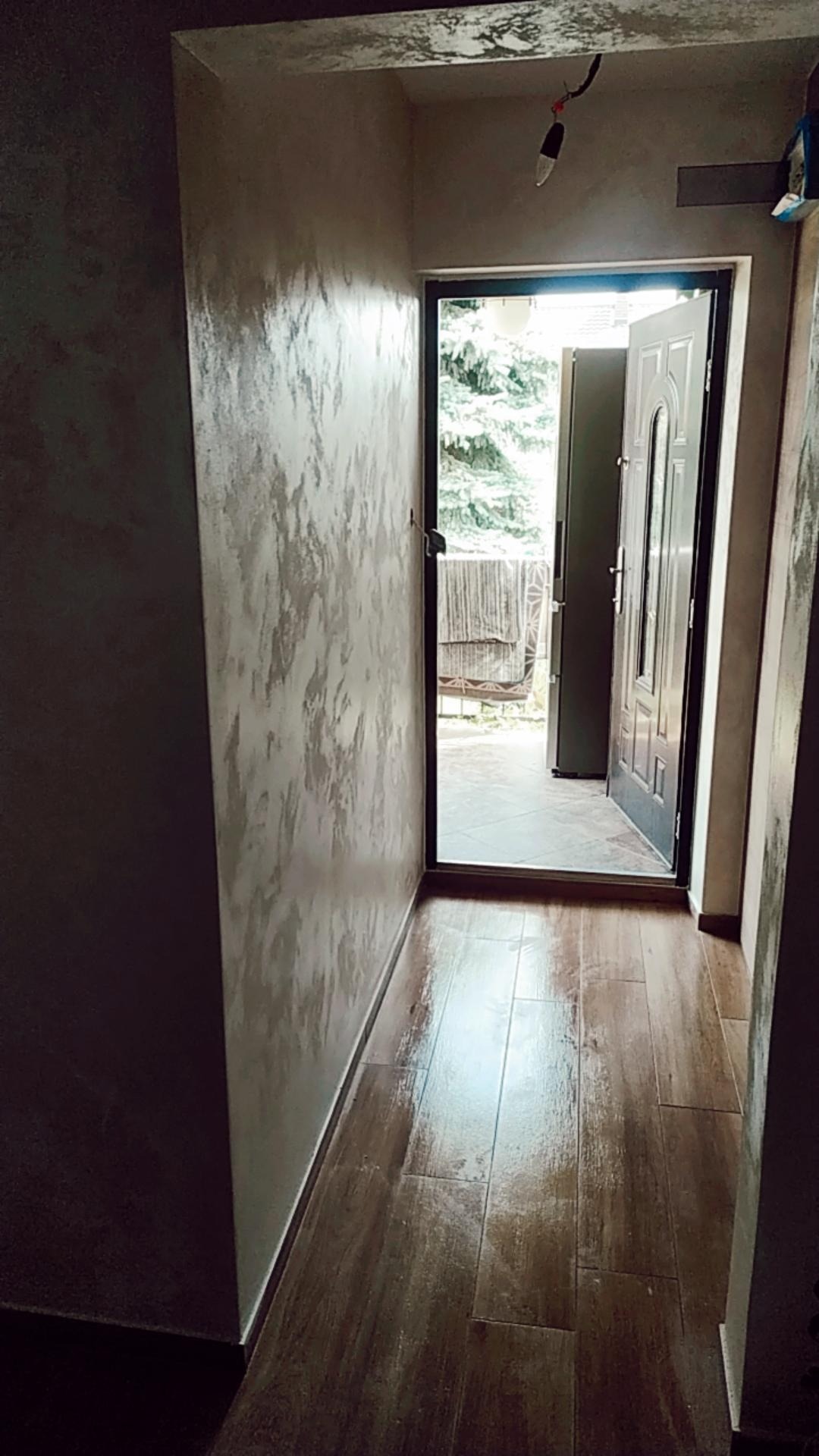 Korytarz z dekoracyjną, szpachlowaną ścianą w odcieniach szarości i beżu, brązowa podłoga imitująca drewno, otwarte drzwi wejściowe z widokiem na zieleń ogrodu.