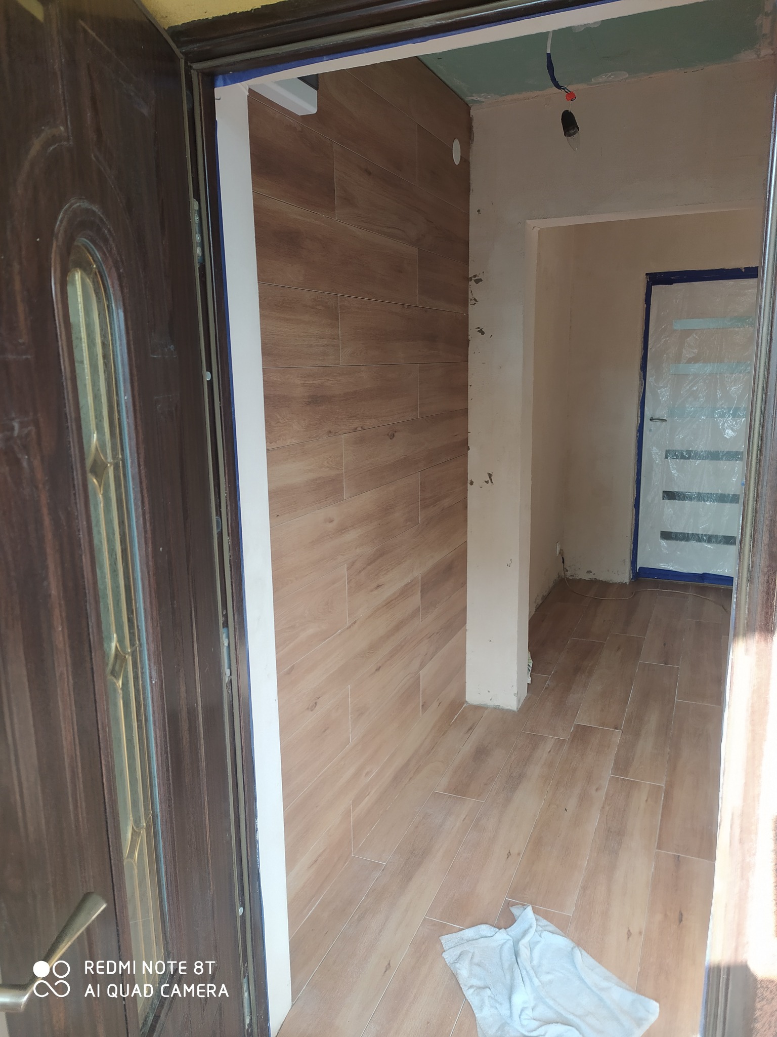 Korytarz w trakcie remontu: ściany i podłoga wyłożone płytkami imitującymi drewno, widoczne niedokończone prace przy suficie i zabezpieczone folią drzwi w głębi.