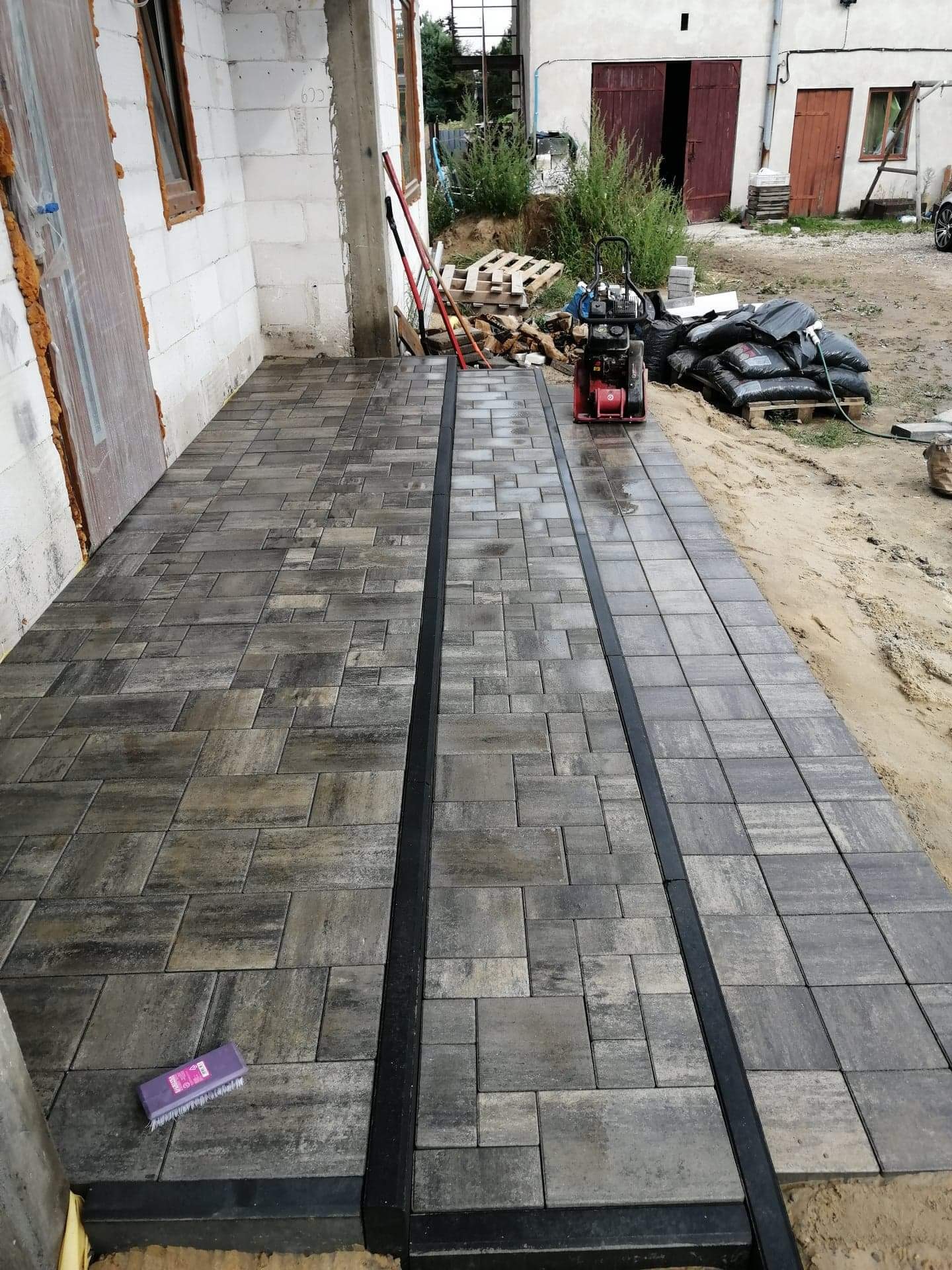 Ułożona kostka brukowa w odcieniach szarości z czarnymi obrzeżami, widoczna ubijarka wibracyjna, budynek w trakcie budowy w tle.