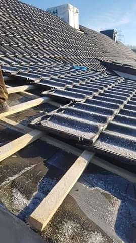 Montaż ciemnej dachówki na konstrukcji dachu z widoczną membraną i łatami, fragment buta pracownika w lewym górnym rogu.