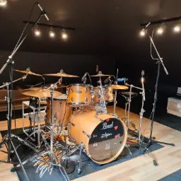 Zestaw perkusyjny Pearl z talerzami Zildjian, mikrofonami i oświetleniem studyjnym w pomieszczeniu z czarnym sufitem i ścianami, na drewnianej podłodze z dywanem.