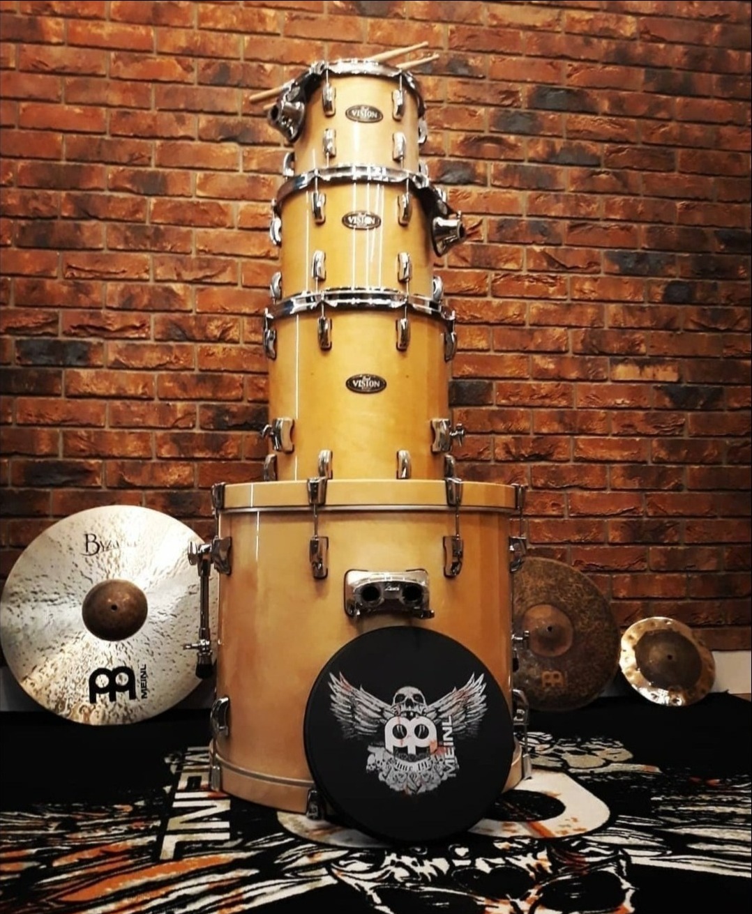 Zestaw perkusyjny marki Vision, ustawiony na tle ceglanej ściany, z talerzami Meinl i tłumikiem z logo Meinl na dywanie z motywem zwierzęcym.