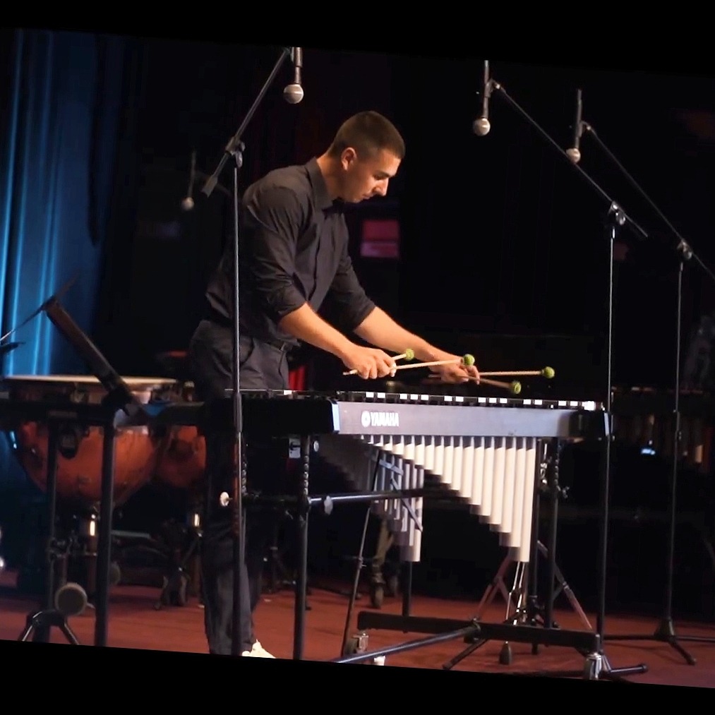 Młody mężczyzna w czarnej koszuli gra na wibrafonie Yamaha z zielonymi pałeczkami na scenie z mikrofonami