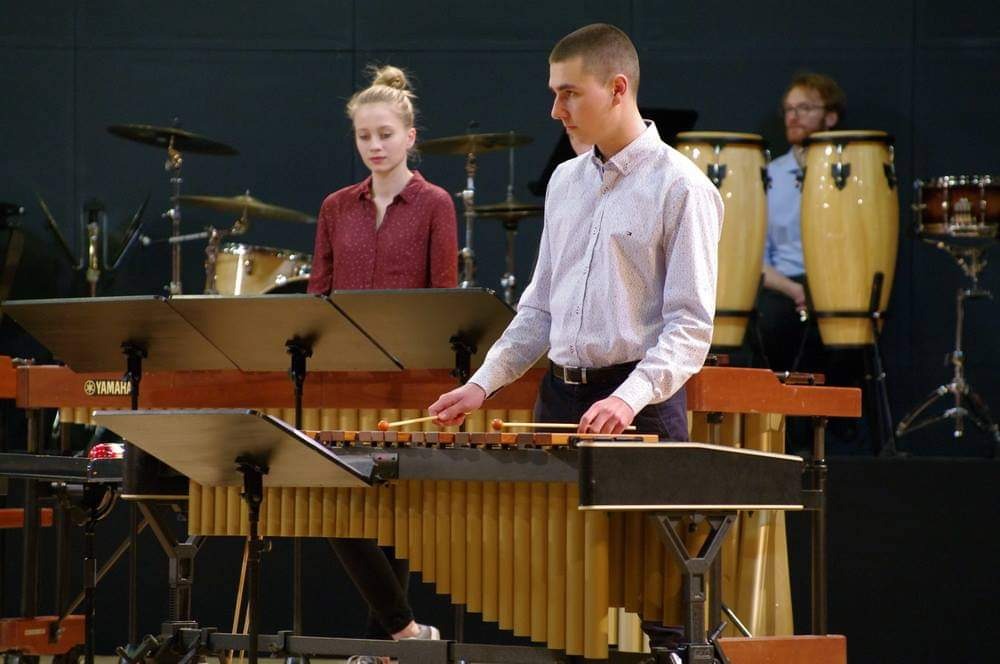 Młody mężczyzna grający na marimbie Yamaha podczas występu, w tle perkusja i inne instrumenty, oświetlenie sceniczne.