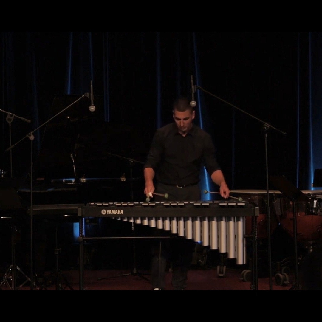 Mężczyzna w czarnej koszuli gra na ksylofonie marki Yamaha na scenie z mikrofonami i fortepianem w tle.