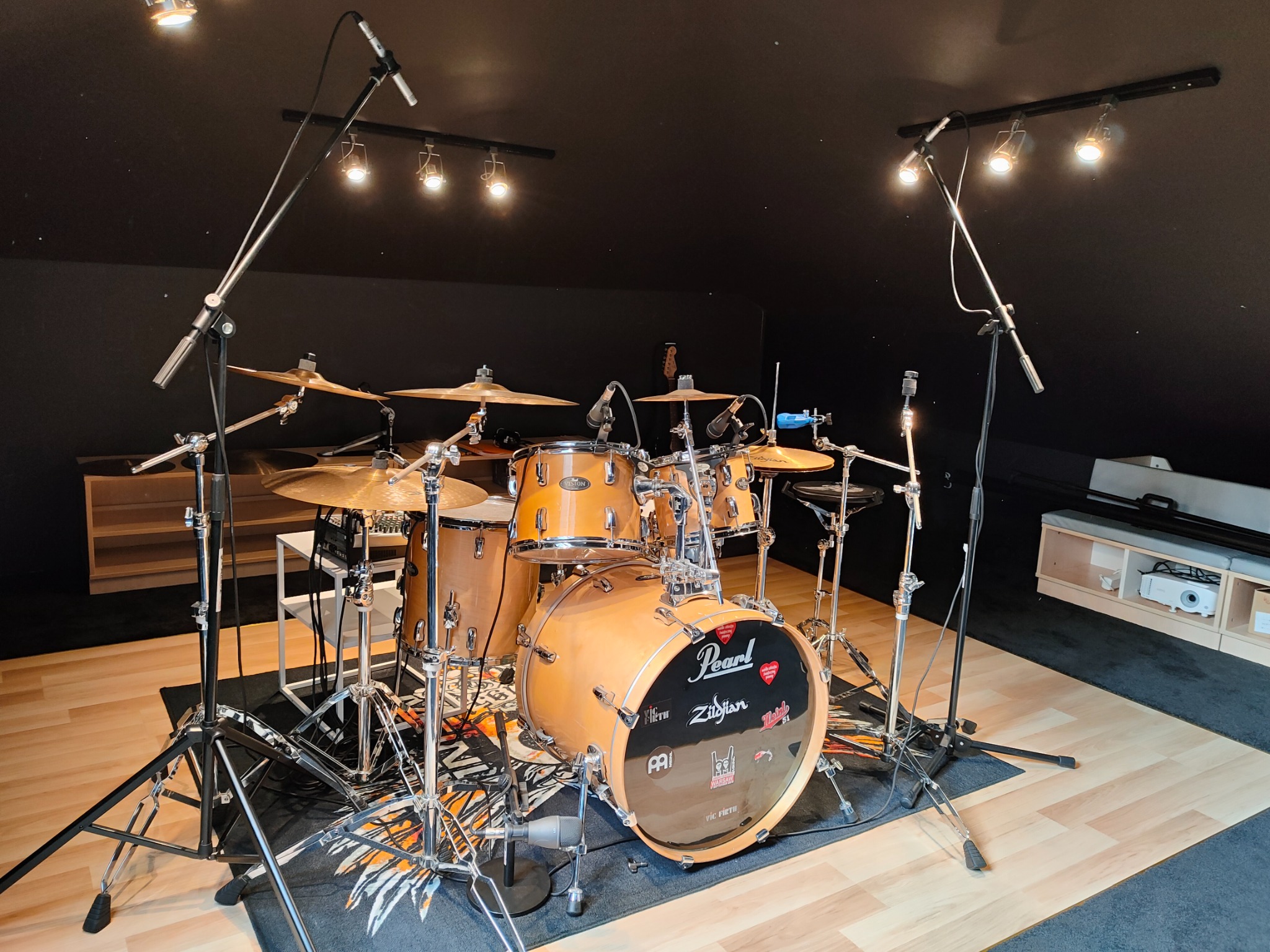Zestaw perkusyjny Pearl z talerzami Zildjian, mikrofonami i oświetleniem studyjnym w pomieszczeniu z czarnym sufitem i ścianami, na drewnianej podłodze z dywanem.