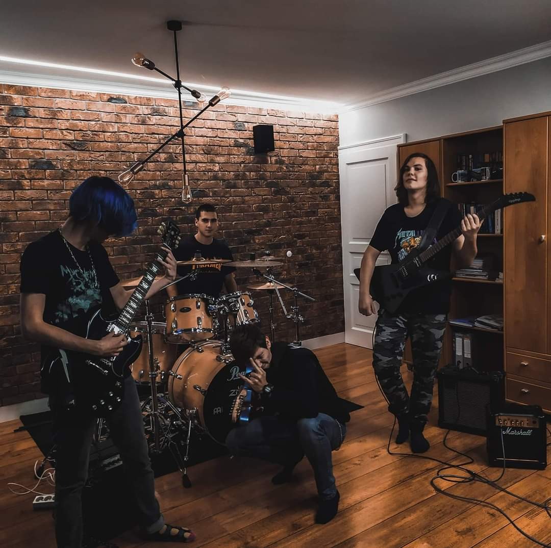 Zespół muzyczny w trakcie próby w pokoju z ceglaną ścianą, perkusista za perkusją, gitarzysta z niebieskimi włosami gra na gitarze, drugi gitarzysta w kamuflażowych spodniach, czwarty muzyk kuca...