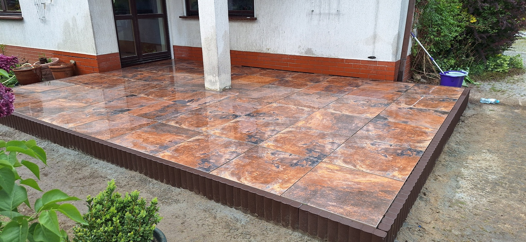 Wykończony taras z płytek imitujących kamień, obrzeże z brązowych elementów, widoczny fragment elewacji budynku z kolumną i roślinność w tle.