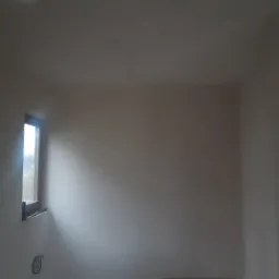 Wnętrze pokoju z oknem po zakończonym nakładaniu gładzi gipsowej na ścianach i suficie. Widoczne gniazdko elektryczne i ślady po instalacjach.