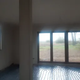 Wnętrze w stanie surowym z widocznym systemem ogrzewania podłogowego w postaci niebieskich rurek ułożonych na folii, duże okna z widokiem na pole i drzewa.