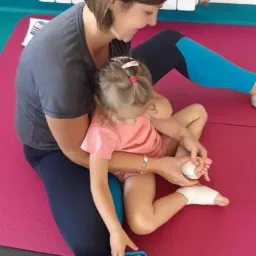 Fizjoterapeutka bandażuje stopę małej dziewczynki siedzącej na różowej macie, z pudełkiem zabawek obok, w jasnym pomieszczeniu z grzejnikiem.